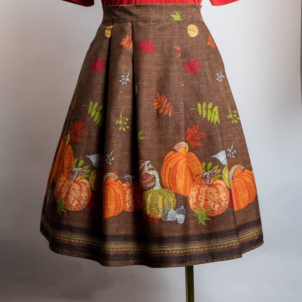 A vintage-style two-piece outfit displayed on a mannequin, featuring a solid bright red short-sleeve mock-neck top and a brown pleated skirt with a colorful autumn print of pumpkins, leaves, and acorns.