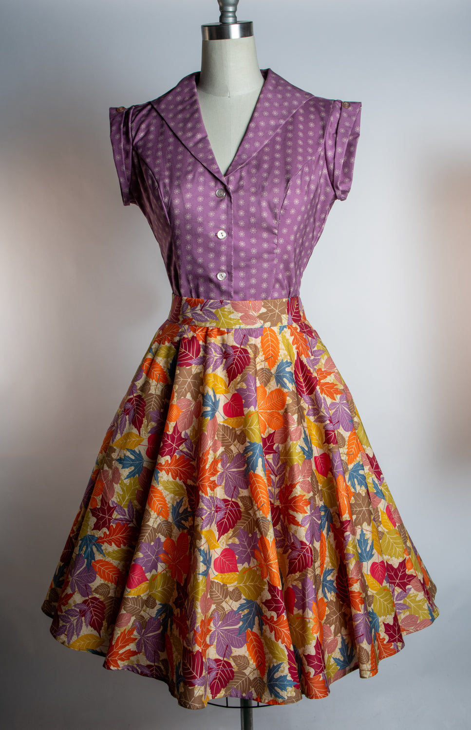 A vintage-style two-piece dress displayed on a mannequin against a plain light gray background. The top is a purple V-neck blouse with white polka dots and short ruffled sleeves.