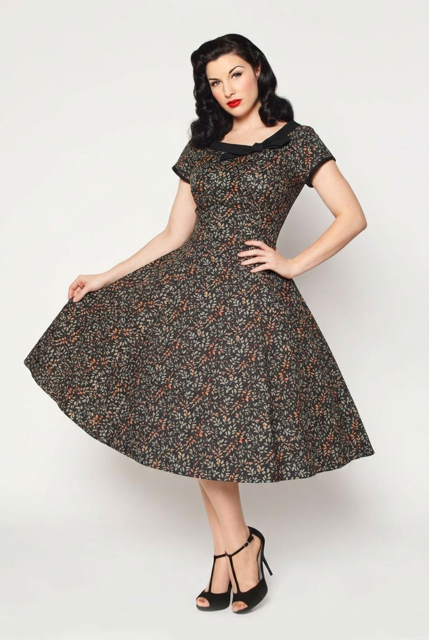 A woman modeling a 1950s-style fit-and-flare dress with a black base and a dense orange and beige floral print. She is posed against a solid light gray studio background, wearing black high-heeled sandals.