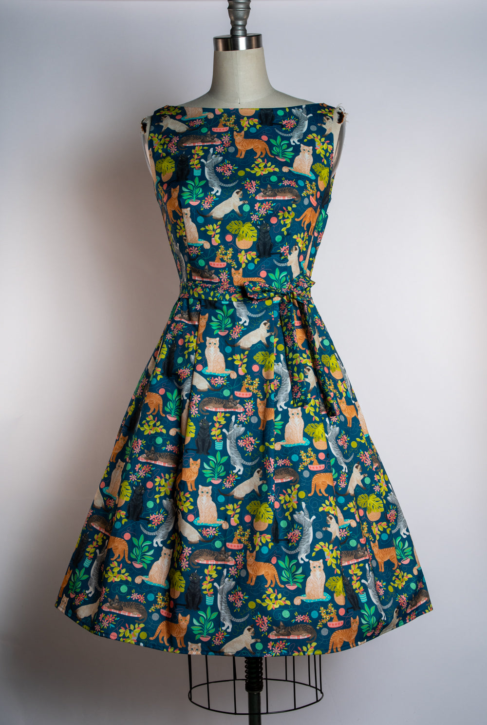 A sleeveless fit-and-flare dress displayed on a mannequin against a plain light gray background. The dress has a dark teal-blue base covered in a whimsical animal print featuring cats, dogs, foxes, rabbits, and foliage in orange, cream, green, and yellow.