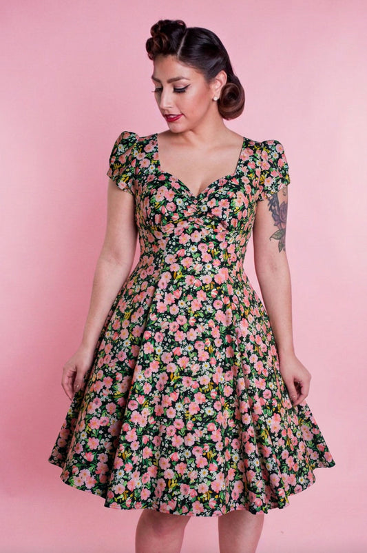 A woman wearing a vintage-style fit-and-flare dress with a pink and green floral print, posed against a solid pink studio background.
