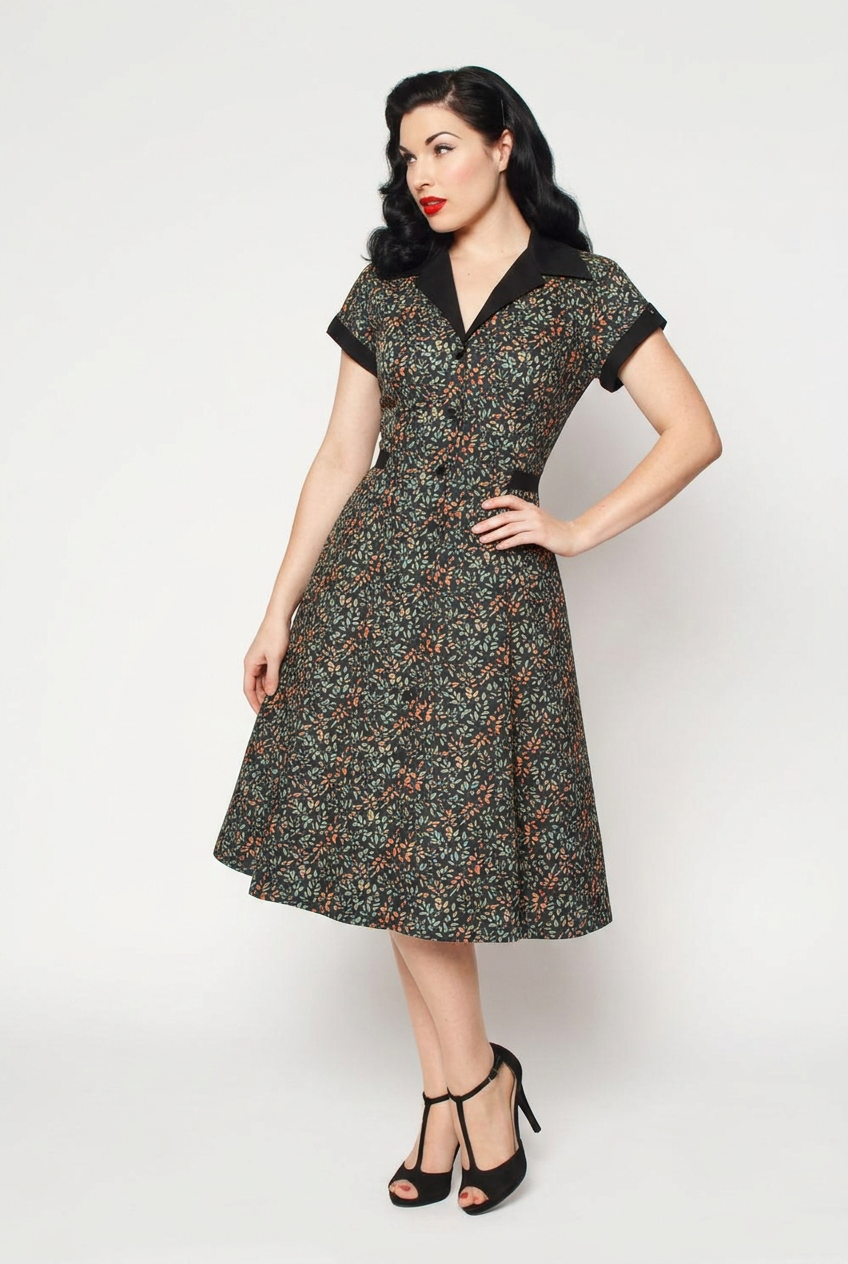 A woman modeling a 1950s-style fit-and-flare dress with a dark green and black floral pattern and black trim, paired with black high heels, against a light gray studio background.