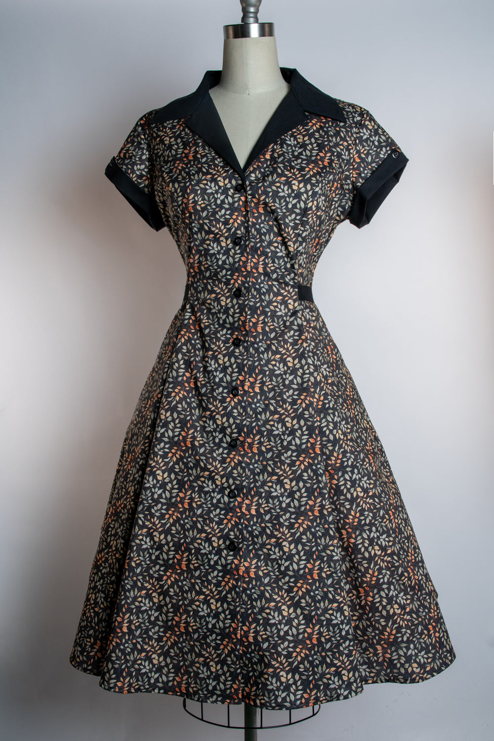 A vintage-style fit-and-flare dress displayed on a mannequin against a plain light gray background. The dress has a black base with a dense small floral print in orange, beige, and white, with solid black short sleeves and a contrasting black collar.