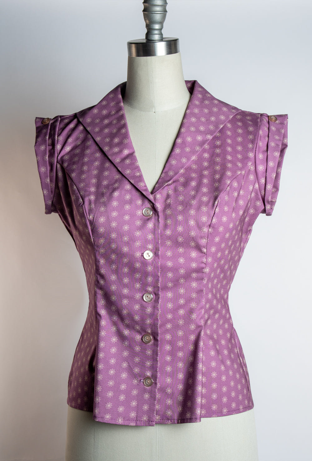 A purple short-sleeve blouse with a white polka dot pattern, displayed on a cream-colored dress form mannequin against a light gray background.