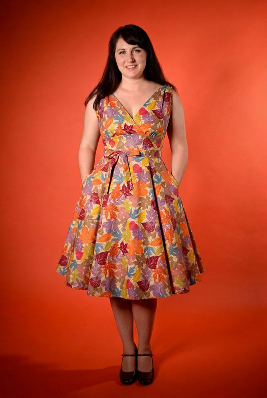 A woman modeling a sleeveless V-neck swing dress with a colorful autumn leaf print, standing against a solid orange-red studio background.