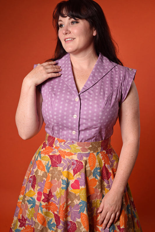 A woman modeling a retro-style two-piece dress with a lavender polka-dot top and a colorful autumn leaf print skirt, set against a solid orange background.