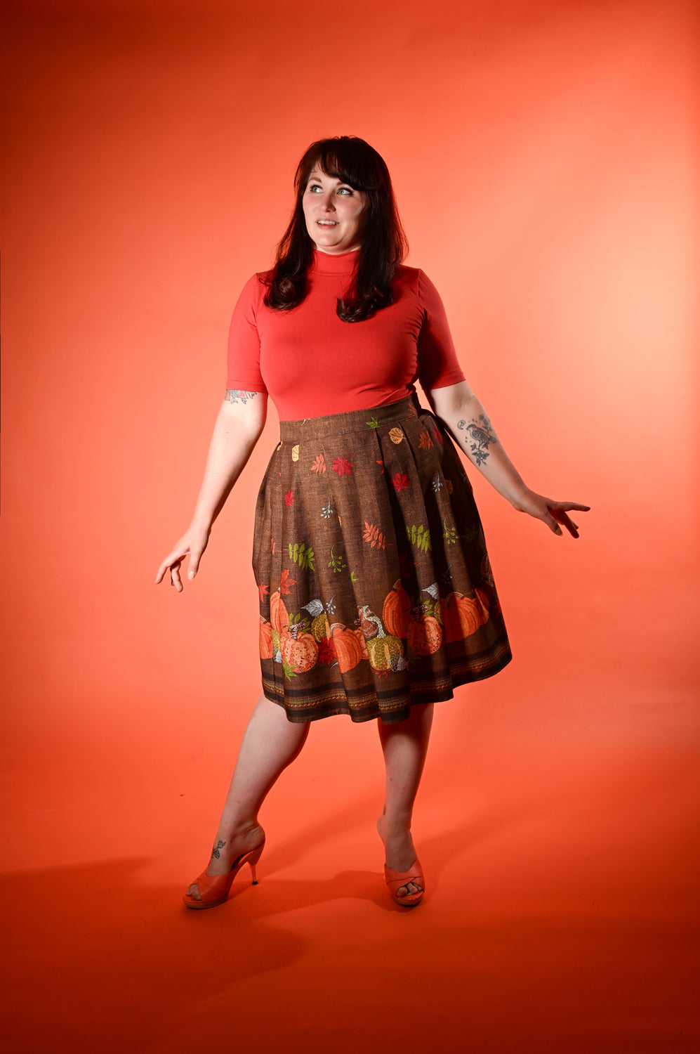A woman modeling a short-sleeve coral red top and a brown A-line skirt with a colorful pumpkin and autumn leaf pattern, posed against a solid orange-red studio backdrop.
