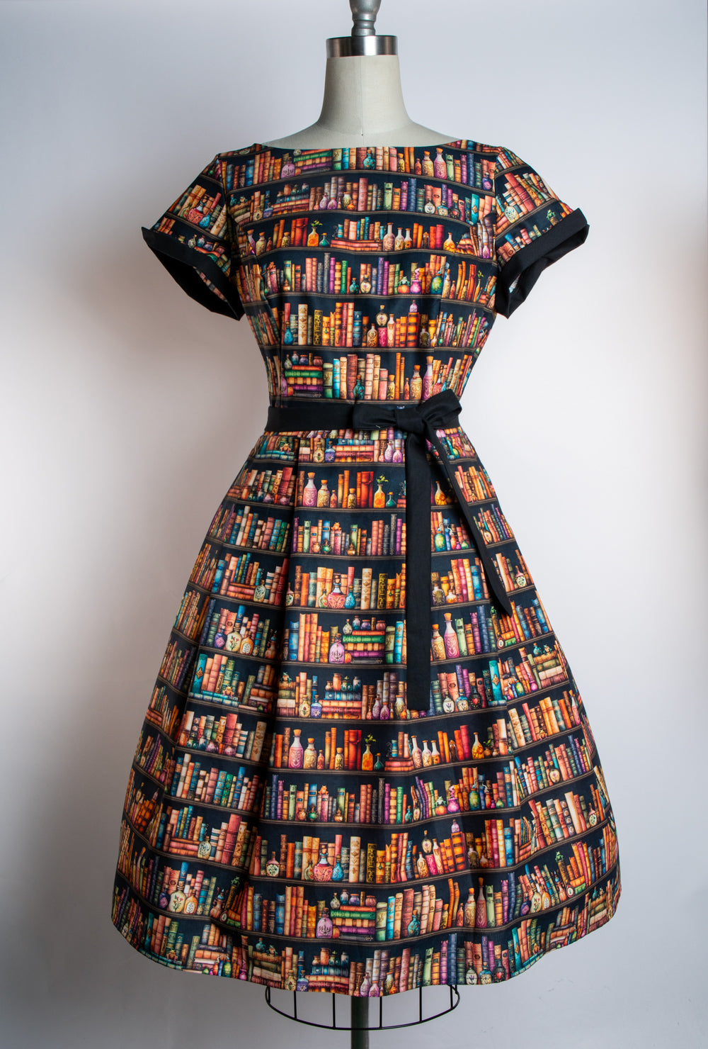 A vintage-style A-line dress displayed on a mannequin, featuring a colorful bookshelf pattern with rows of book spines in various colors on a dark background, with a black waist tie belt.
