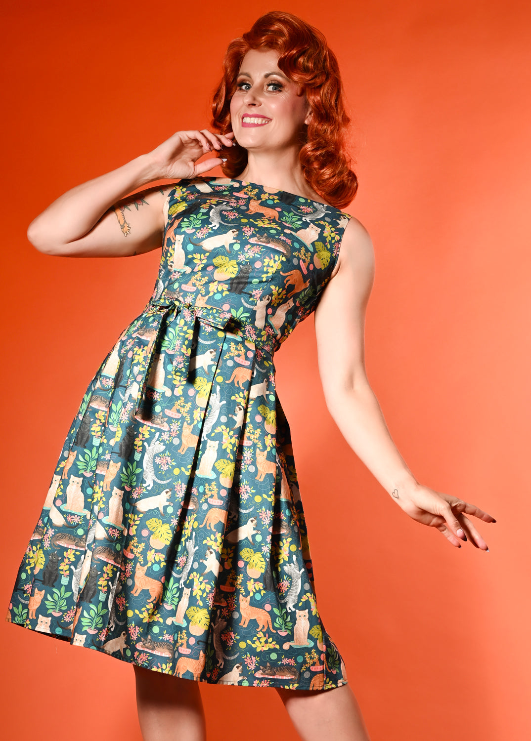 A woman modeling a sleeveless fit-and-flare dress with a colorful animal print on a teal background, posed against a solid orange backdrop.
