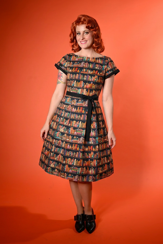 A woman with red hair wearing a retro-style A-line dress with a colorful bookshelf pattern and black trim, standing against a solid orange background.