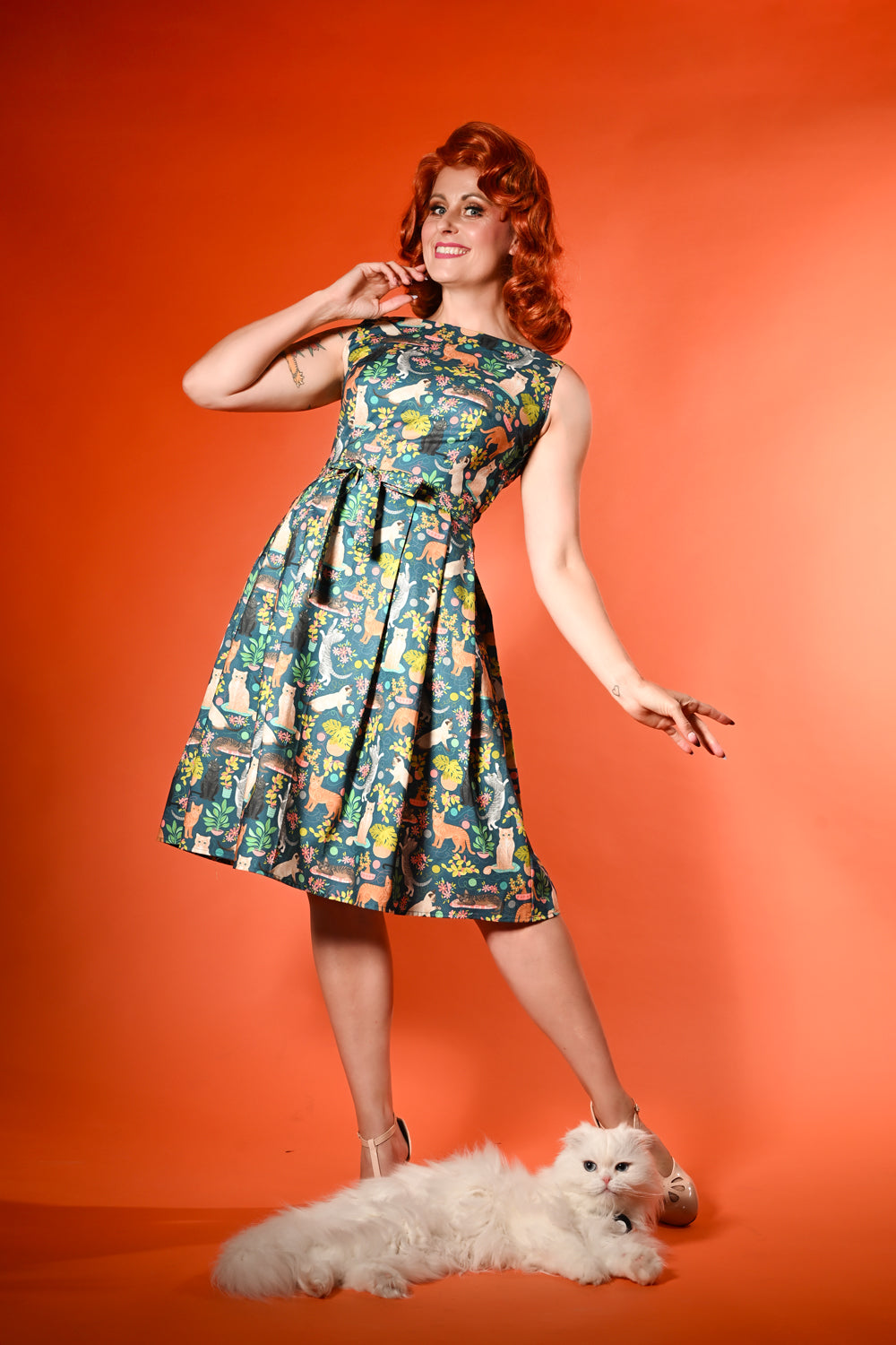 A woman with red hair poses in a sleeveless, knee-length A-line dress with a colorful retro-style cartoon print, standing against a solid orange background with a small white dog at her feet.