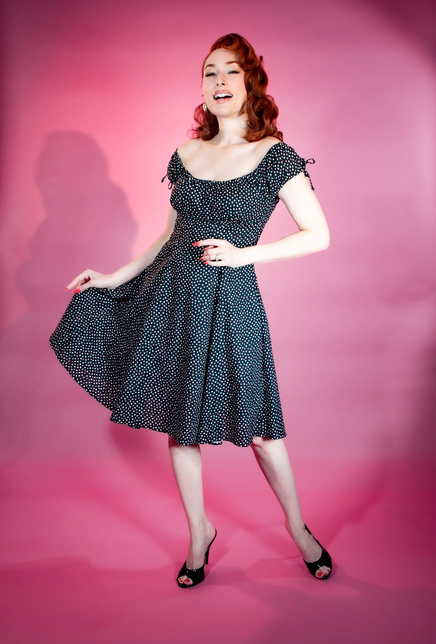 Woman in a black polka dot dress posing against a pink background